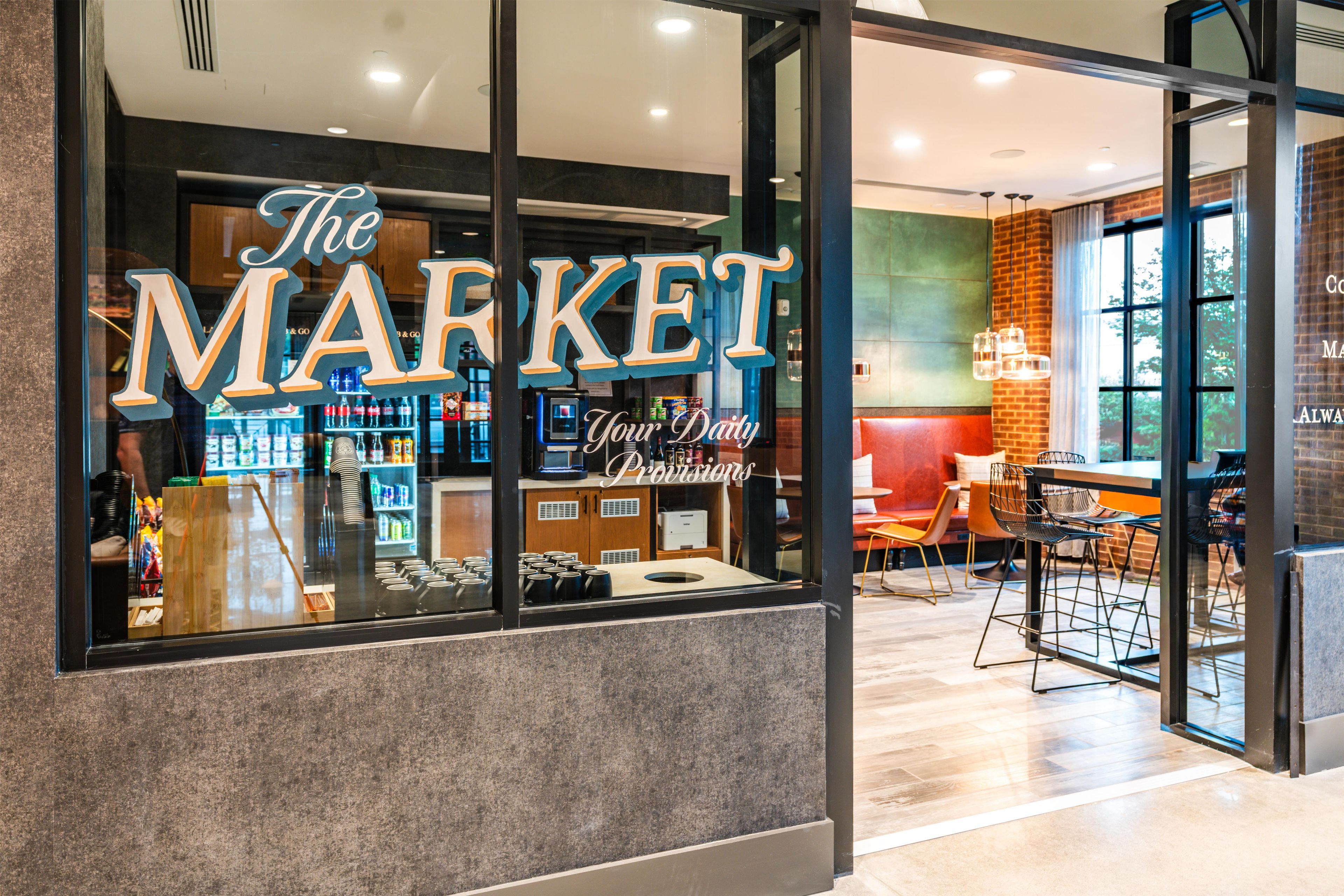 Una vetrina con un grande cartello che recita 'The MARKET Your Daily Provisions'. All'interno, scaffali pieni di prodotti, un'area con tavoli e sedie, e un bancone.