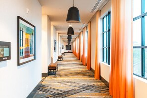 A hotel corridor with a patterned carpet, benches, framed artwork, and large windows with orange curtains.