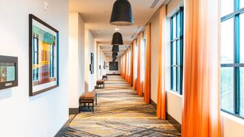 Un pasillo de hotel con alfombra estampada, bancos, obras de arte enmarcadas y ventanas amplias con cortinas naranjas.