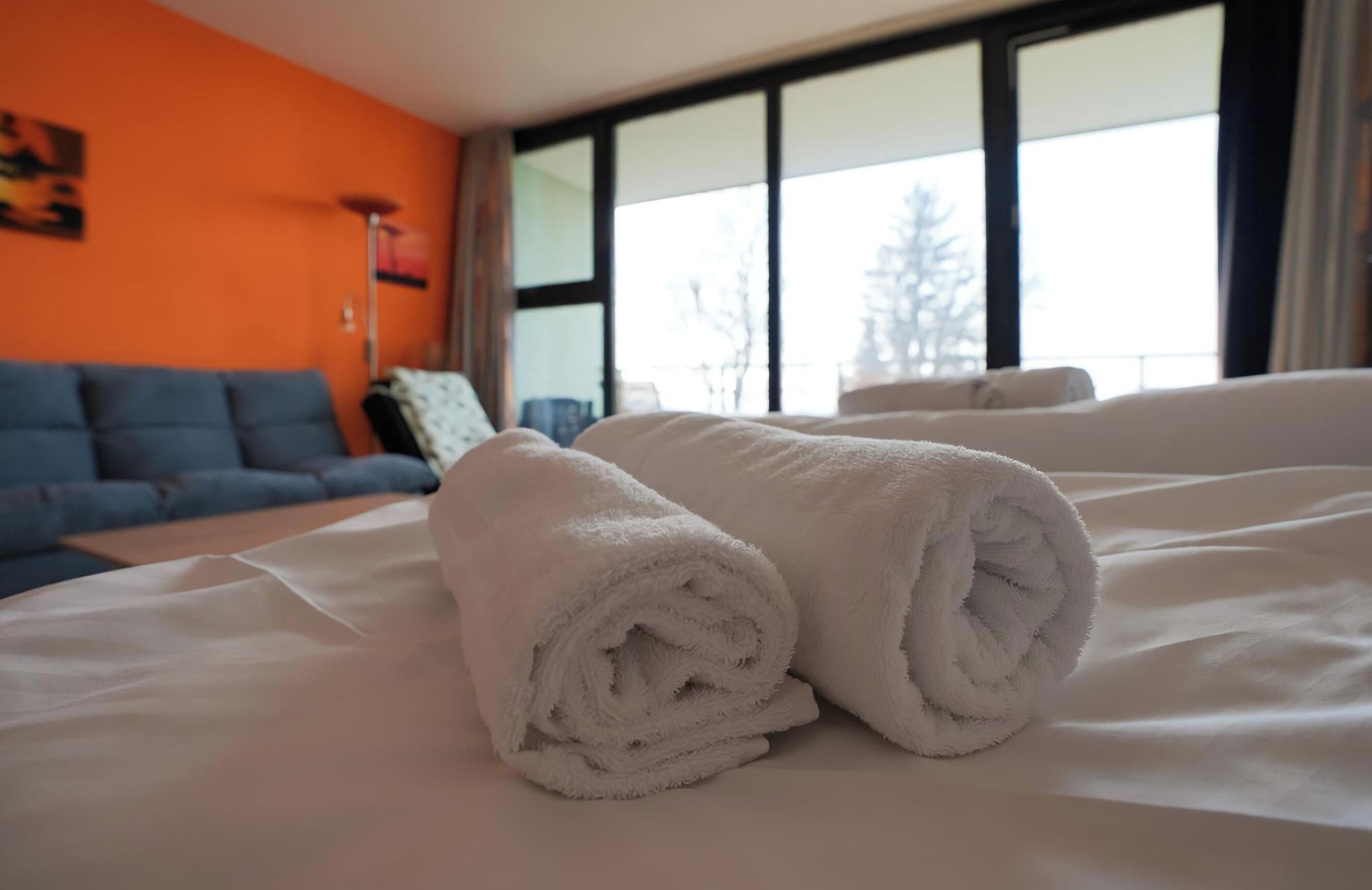 Una cama bien tendida con toallas blancas enrolladas, un sofá azul y un ventanal con vistas a árboles.
