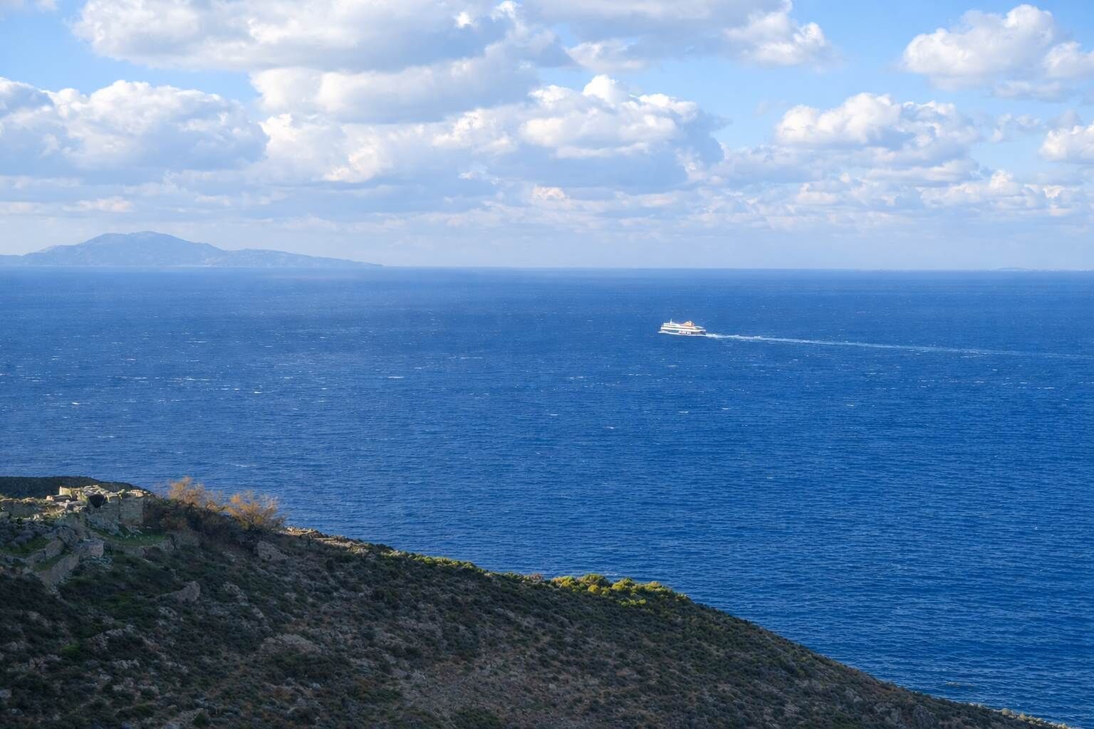 Paisatge costaner amb un vaixell al mar, terreny rocosós i muntanyes a la distància.