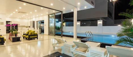 A modern indoor pool area with a glass enclosure, white lounge chairs, and a staircase leading to a second level.