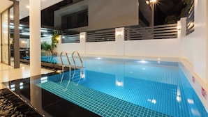 Une piscine intérieure aux carreaux bleu clair, avec un escalier en métal et un mur blanc orné d’un luminaire horizontal.