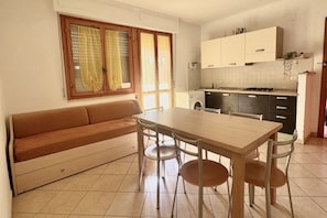 A compact living space with a sofa, dining table, chairs, a kitchenette, and a window with curtains. - Staying at the Marina di Bibbona Apartments offers the perfect blend of home away from home convenience and the comfort of a holiday immersed in nature, just a short walk from the sea and all essential services.With its inviting atmosphere, Marina di B (Marina di Bibbona)