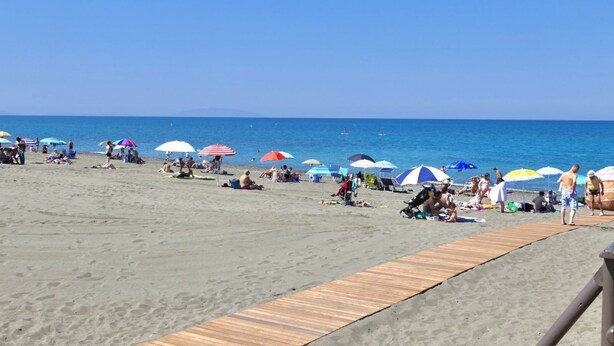 Una playa con gente relajándose bajo sombrillas coloridas, un paseo marítimo de madera y el océano al fondo.