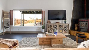 A living room with a large TV, wooden furniture, a fireplace, and a view of the outdoors. - Pueblo Villa - Sleeps 12 • 2 Homes on 2.5 Acres (Joshua Tree)