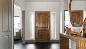 A hallway with a wooden door, a wicker console table, and a bed in the background. - Pueblo Villa - Sleeps 12 • 2 Homes on 2.5 Acres (Joshua Tree)