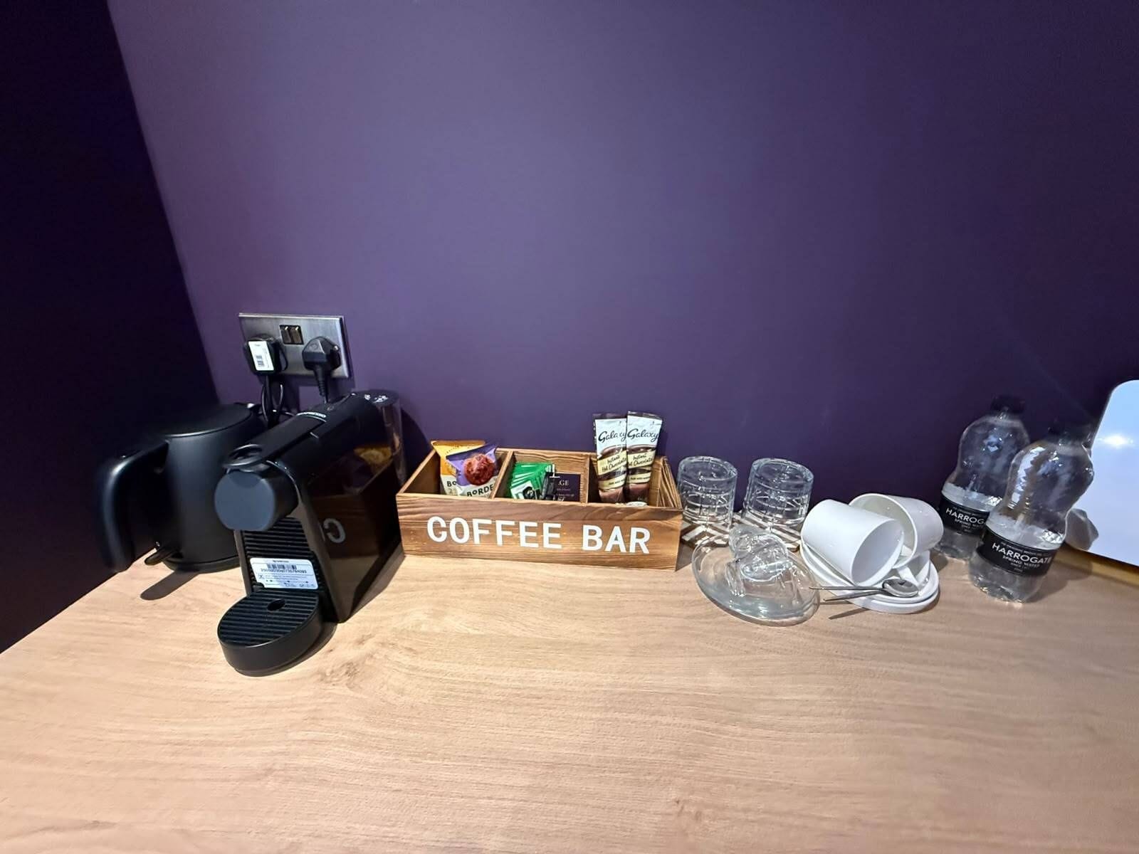 A coffee bar setup with a coffee machine, a wooden box labeled 'COFFEE BAR,' coffee pods, glasses, and a water dispenser.