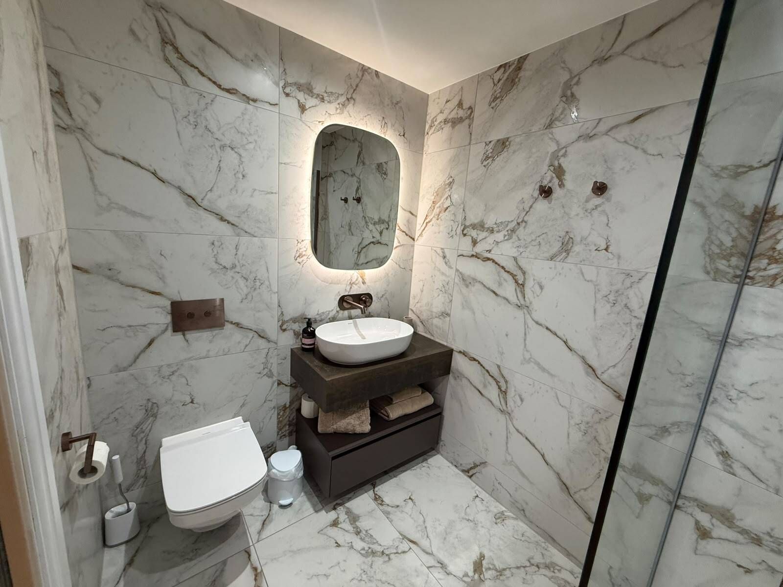 A bathroom with a marble wall, a round mirror, a sink, a toilet, and a shower area.