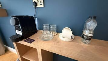 A room with a wooden desk, a black coffee machine, three glasses, a white plate, and a water bottle.