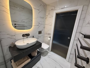 A modern bathroom with a white sink, a mirror with under-cabinet lighting, a toilet, and a shower area. - Auberge (Lytham St. Anne's)