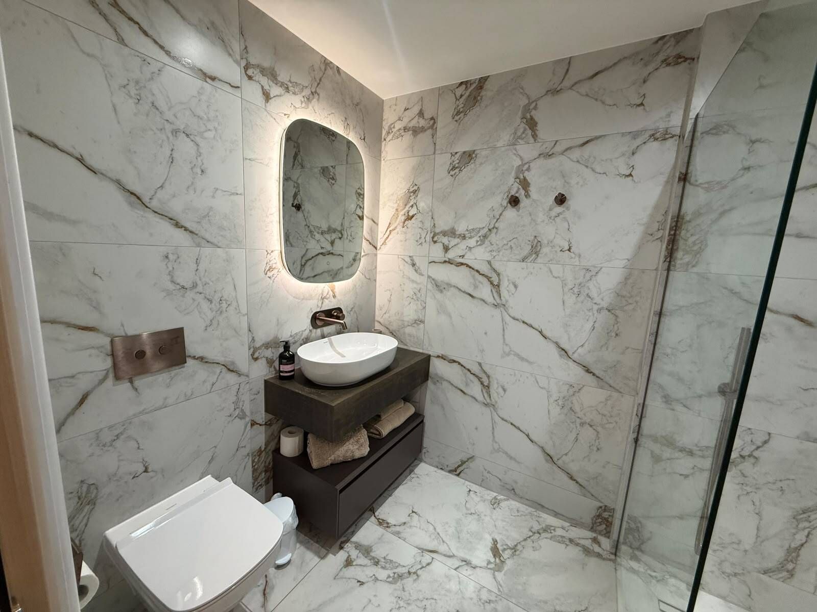 A bathroom with a marble wall, a round sink, a mirror, a toilet, and a shower area.