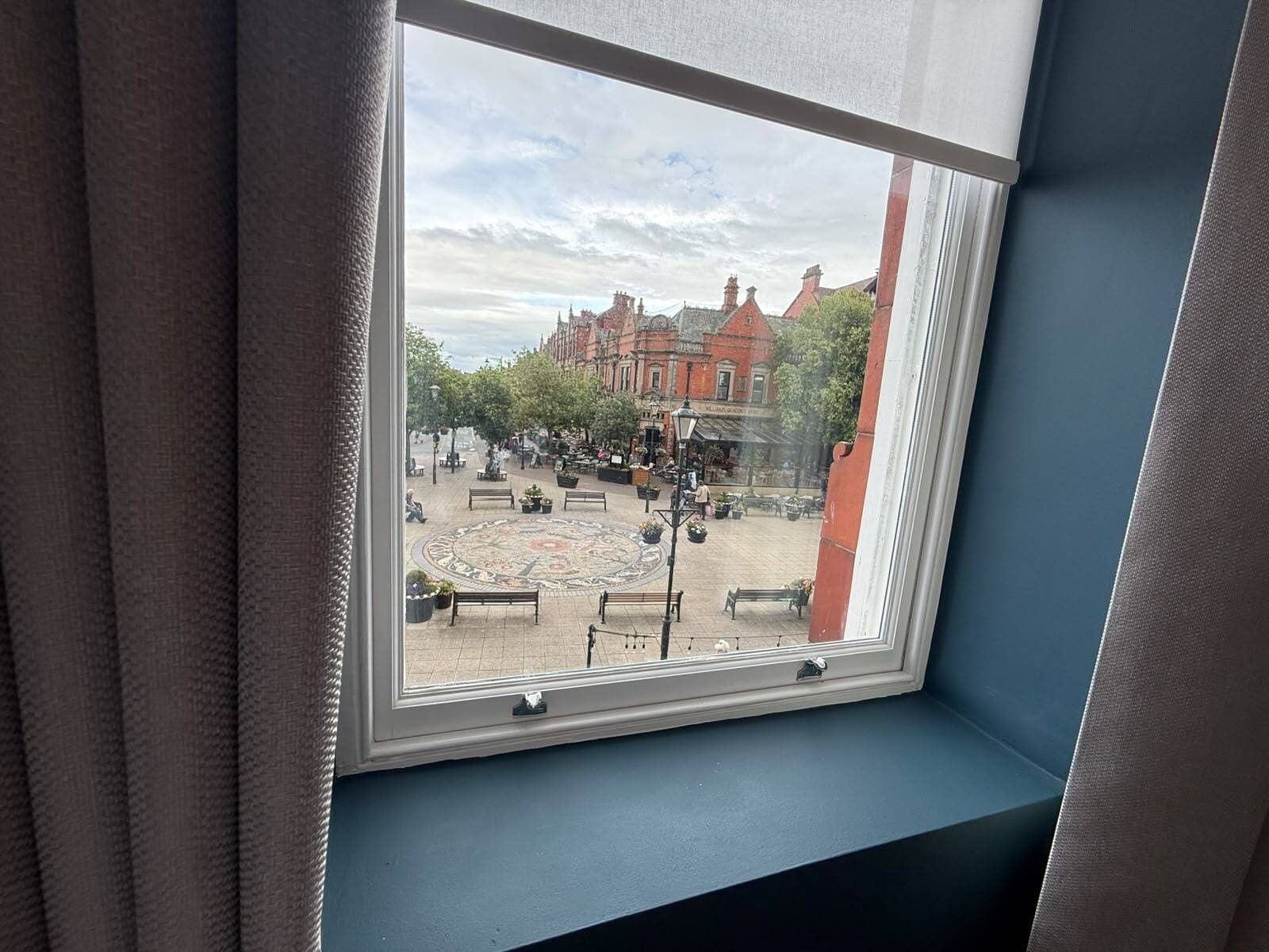 A window with a view of a courtyard with benches, a patterned pavement, and red brick buildings.
