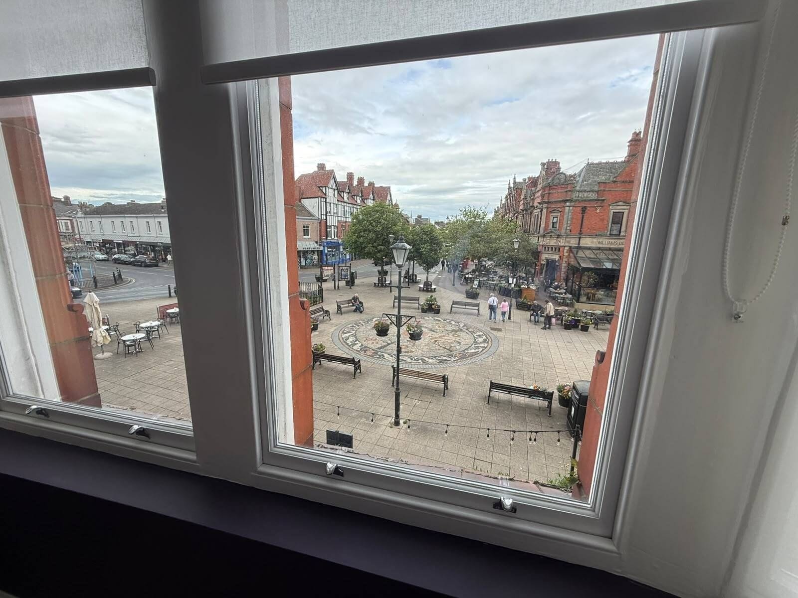 A view through a window of a public square with benches, a circular flower bed, and buildings in the background.