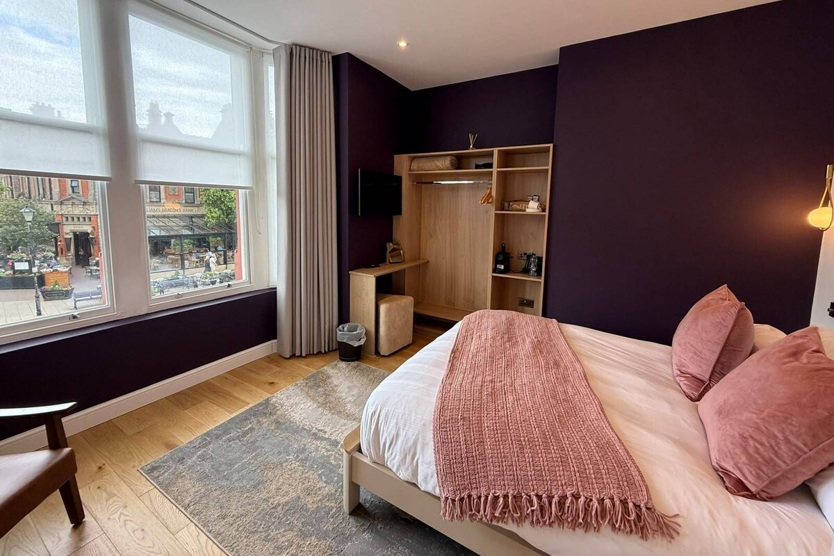 A bedroom with a bed, a nightstand, a wardrobe, a window with a view of the street, and a small table.