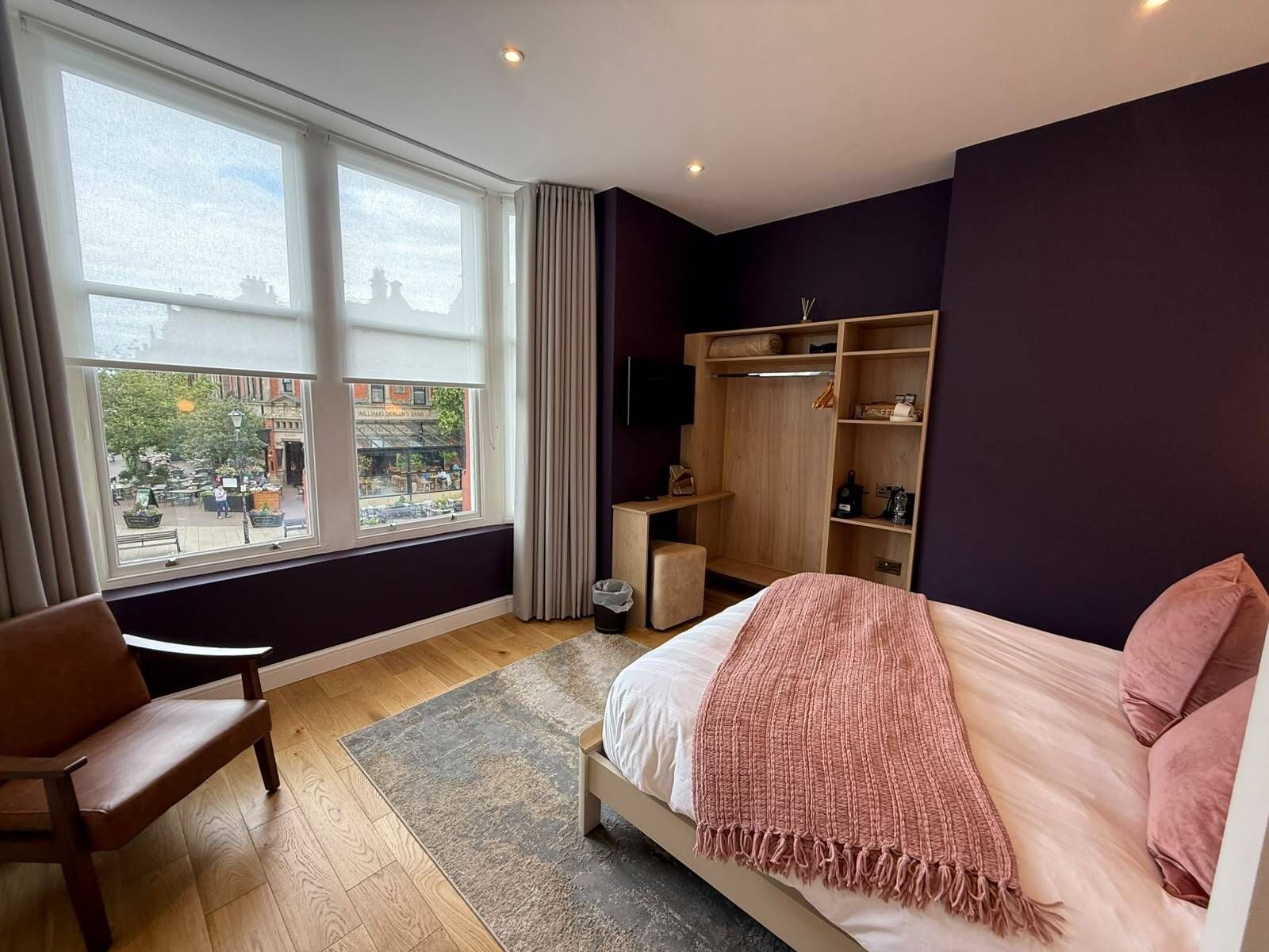 A bedroom with a bed, a chair, a wooden wardrobe, and a window with a view of the street.