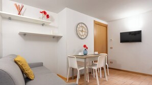 Uma sala de estar com um sofá cinza, uma mesinha com cadeiras, uma televisão na parede e uma porta de madeira.