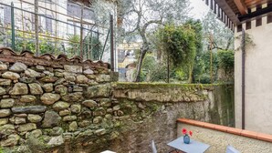 Um muro de pedra, uma pequena mesa com cadeiras e vista para um jardim com árvores e um edifício ao fundo.