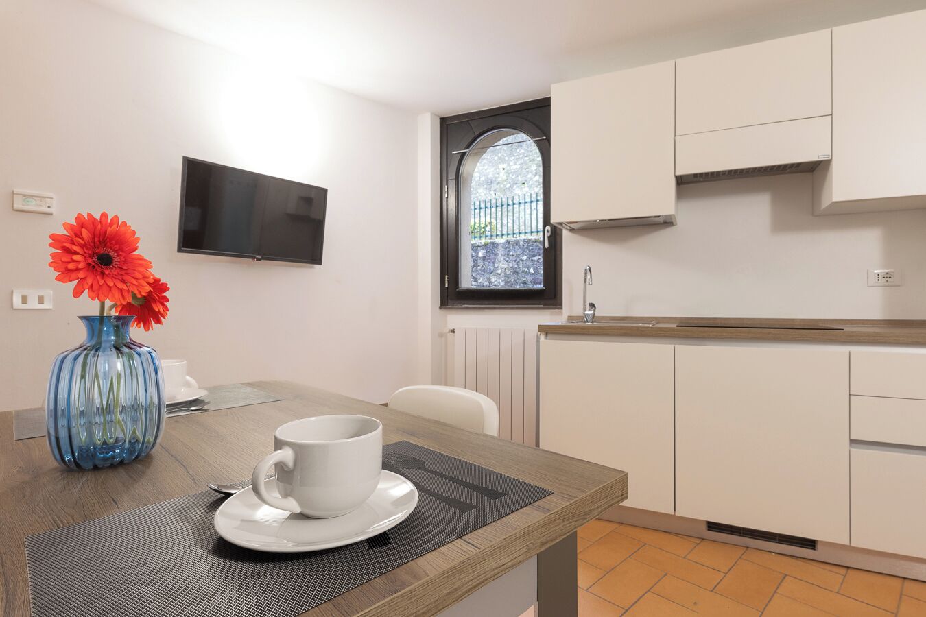 A small kitchenette with a sink, cabinets, and a window. There is a table with a vase and a cup.