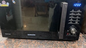 A black Samsung smart oven with a digital display, control buttons, and a power dial. - Diamond Villa (Gautam Buddh Nagar)