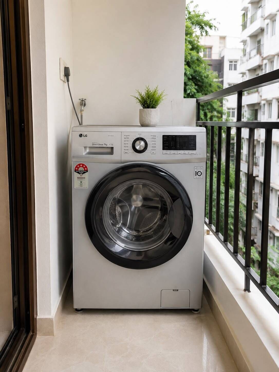 A front-loading LG Direct Drive 7kg washing machine on a balcony with a small potted plant on top, a power outlet, and a view of a residential building.