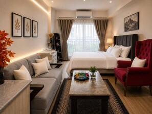 A hotel room with a bed, a red armchair, a grey sofa, a coffee table with a tray of glasses, and a bookshelf.