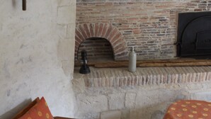 Une cheminée en pierre surmontée d’une arche en briques, un canapé en bois avec des coussins à motifs et une table recouverte d’une nappe à motifs.