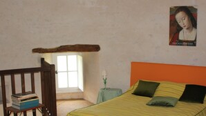 Une chambre à coucher avec un lit, une fenêtre, une petite table avec une lampe et une pile de livres sur une chaise.