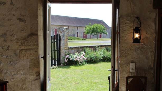 Une porte ouverte donne sur une cour verdoyante, avec un bâtiment visible au loin.