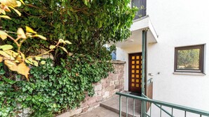 Una escalera conduce a una puerta con diseño de cuadrícula, rodeada de follaje y un edificio blanco con una ventana.