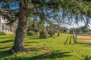 A grassy area with a large tree, a fenced area, and a building with windows in the background. - Alloggio turistico Isola dei Cavalli (Manziana)