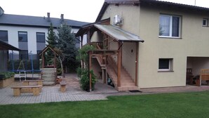 Un edificio residenziale con un portico coperto, una scala che conduce al secondo piano e un piccolo giardino con una panchina e un’area giochi.