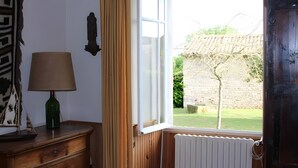 An open window with a view of a garden, a wooden dresser with a lamp, and a wall-mounted decoration.