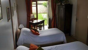 A hotel room with two beds, a desk with a chair, a small table, and a view of the garden through an open door.