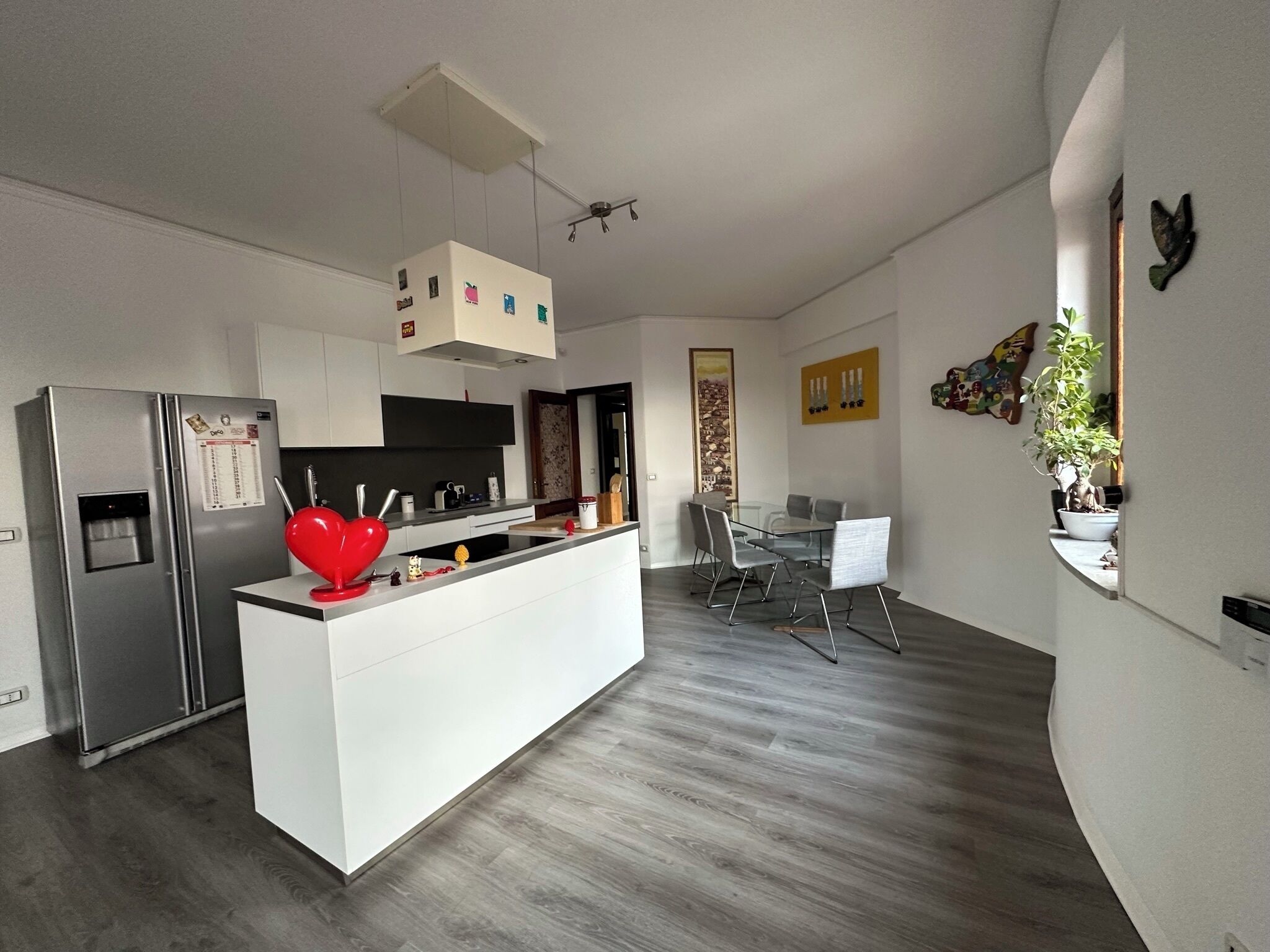 Una cucina moderna con isola centrale, zona pranzo con tavolo in vetro e angolo living con divano e televisione.