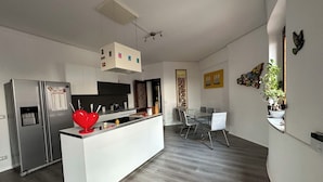 Una cocina moderna con una isla central, un comedor con mesa de vidrio y un espacio de sala de estar con sofá y televisor.
