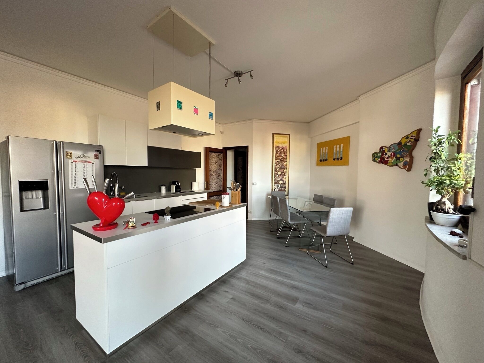 Una cucina moderna con isola centrale, zona pranzo con tavolo in vetro e angolo living con una pianta.