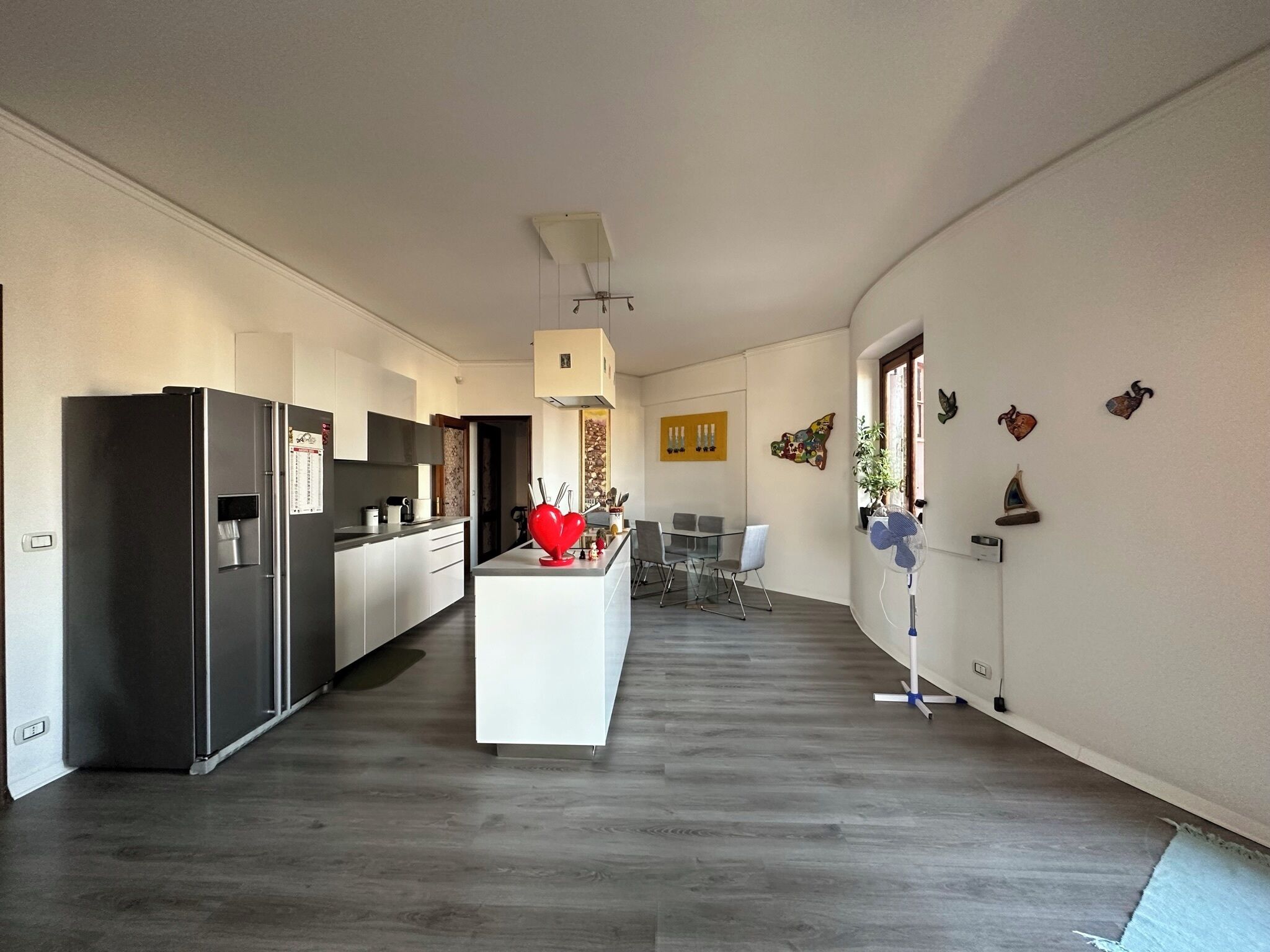 Una cucina moderna con isola centrale, una zona pranzo con tavolo e sedie, e un corridoio che conduce a una camera da letto.