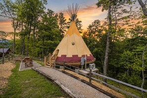 Ett tipi-tält med en träveranda och en bänk, omgivet av träd och en solnedgång.