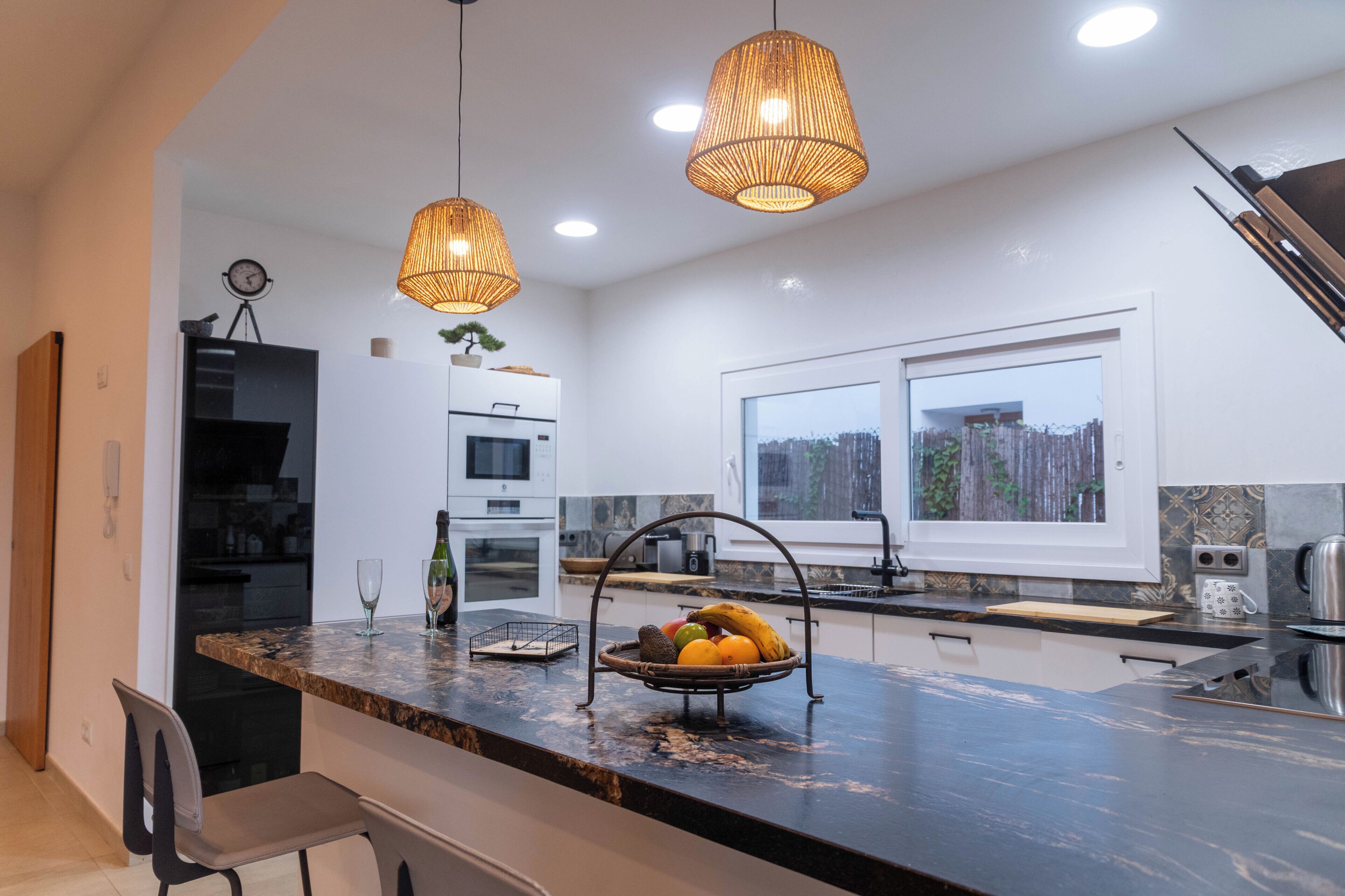 A modern kitchen with a marble countertop, pendant lighting, and a fruit basket.