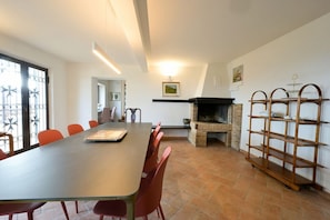 Un espace repas comprenant une longue table, des chaises rouges, une cheminée et une étagère en bois.