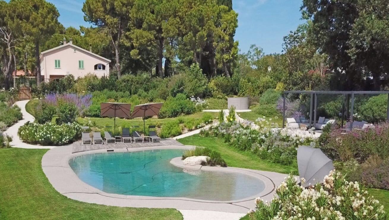A swimming pool with a curved edge, surrounded by a well-maintained lawn and various plants. There is a house in the background, and a gazebo with a roof.