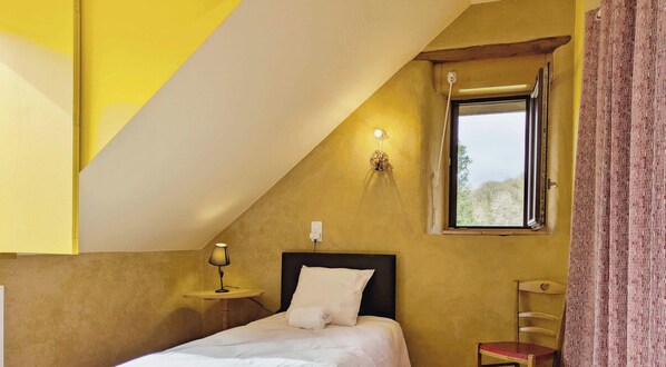 Quarto com teto inclinado, cama com roupa de cama branca, mesa de cabeceira com abajur e janela com vista para árvores.