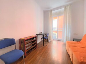 Une pièce avec un plancher en bois, une chaise bleue, un canapé orange, une petite table avec des chaises et une fenêtre avec des rideaux.