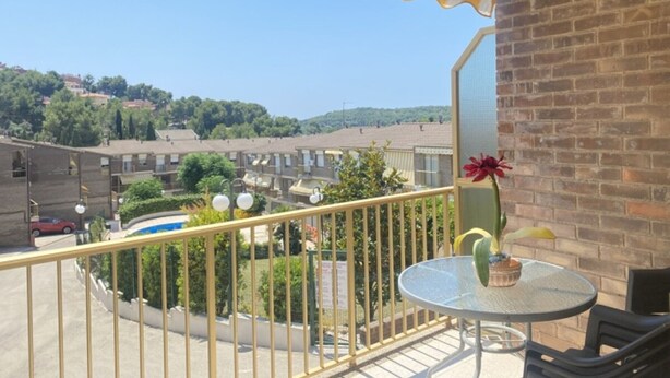 A balcony with a yellow and white striped awning, a round glass table with a potted plant, and a view of a residential area with brick buildings and greenery. - House in La Mora - 8 guests (Tarragona)