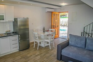 Eine kleine Küche mit Kühlschrank, Esstisch und Stühlen, ein graues Sofa und eine Holztür, die nach draußen führt.