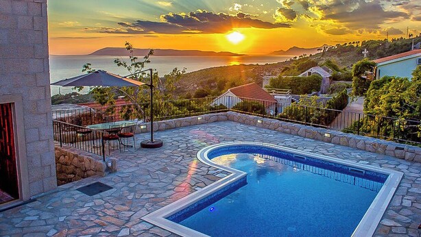 En swimmingpool med stenterrasse, en patio med udendørs møbler og havudsigt i solnedgangen.