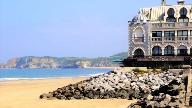 Un hotel frente al mar con fachada de piedra, vistas a la playa y un litoral rocoso.