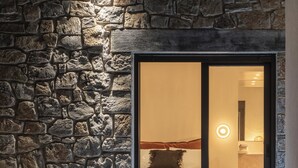 Un mur en pierre, une chaise et une porte-fenêtre coulissante donnant sur une pièce avec un lit et un luminaire circulaire.