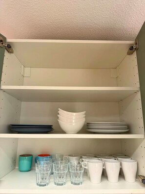 Una repisa de cocina con cuencos apilados, platos y una hilera de vasos de vidrio y tazas blancas.